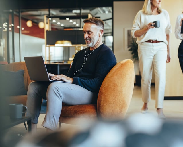 Une personne est assise sur une chaise orange, travaillant sur un ordinateur portable avec des écouteurs, tandis que deux autres se tiennent à proximité, l'une d'elles tenant une tasse.