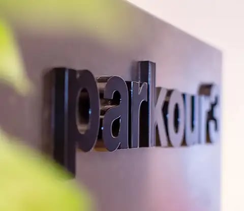 Logo noir de Parkour3 en trois dimensions plaqué sur une surface lisse, partiellement masqué par une plante verte.