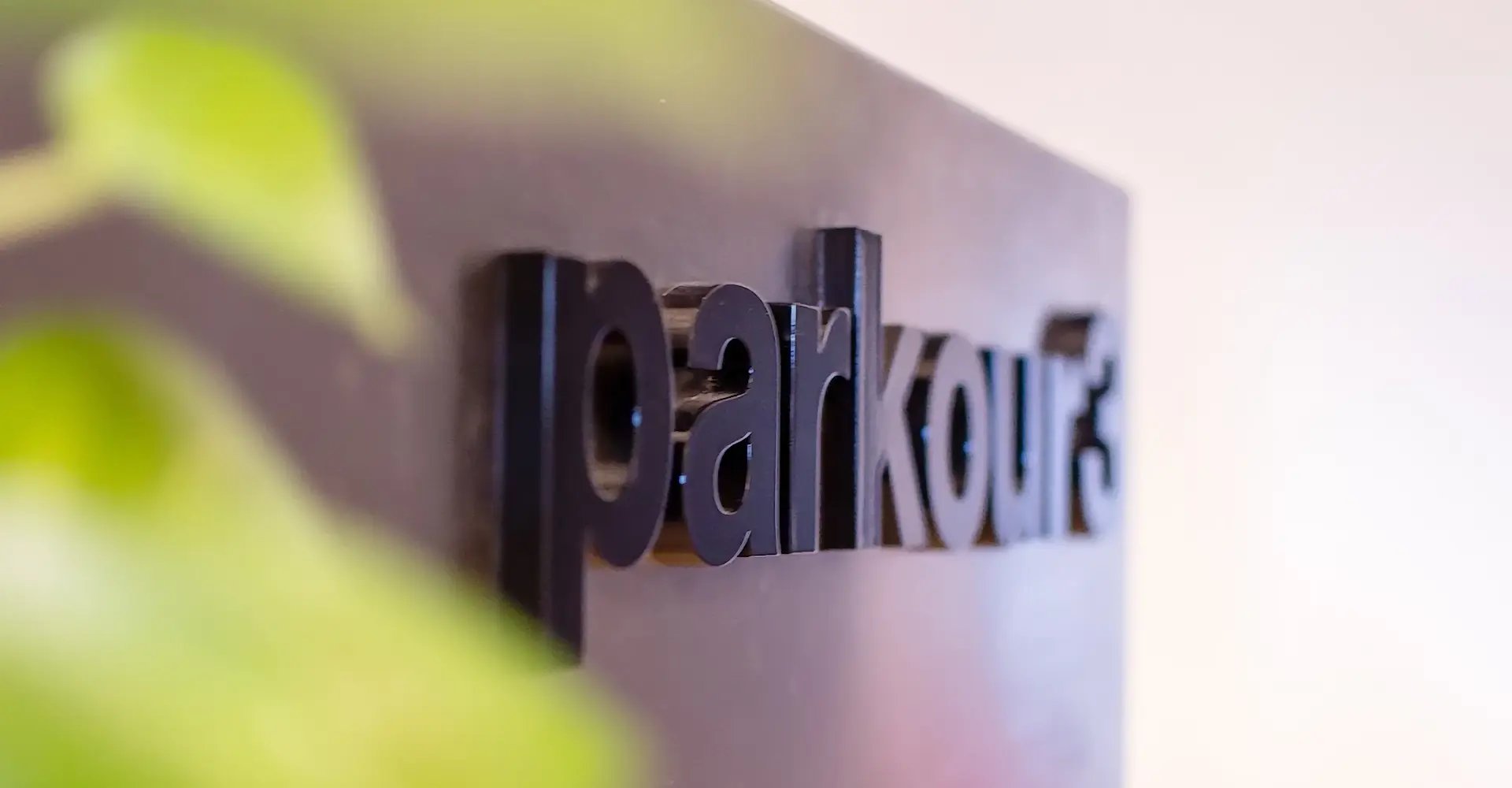 Three-dimensional black Parkour3 logo on a smooth surface, partially obscured by a green plant.