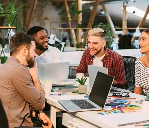 Un groupe de personnes diverses collabore de manière très amicale autour d'une table remplie d'ordinateurs portables, de cahiers et de plantes dans un espace de travail moderne et lumineux.