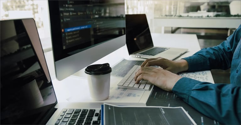 A person typing on a keyboard at a desk equipped with several computers, a cup of coffee and documents showing coding and design diagrams.