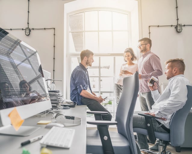 A professional group discussion in a bright office, with four people engaged in conversation near a desk and computer.