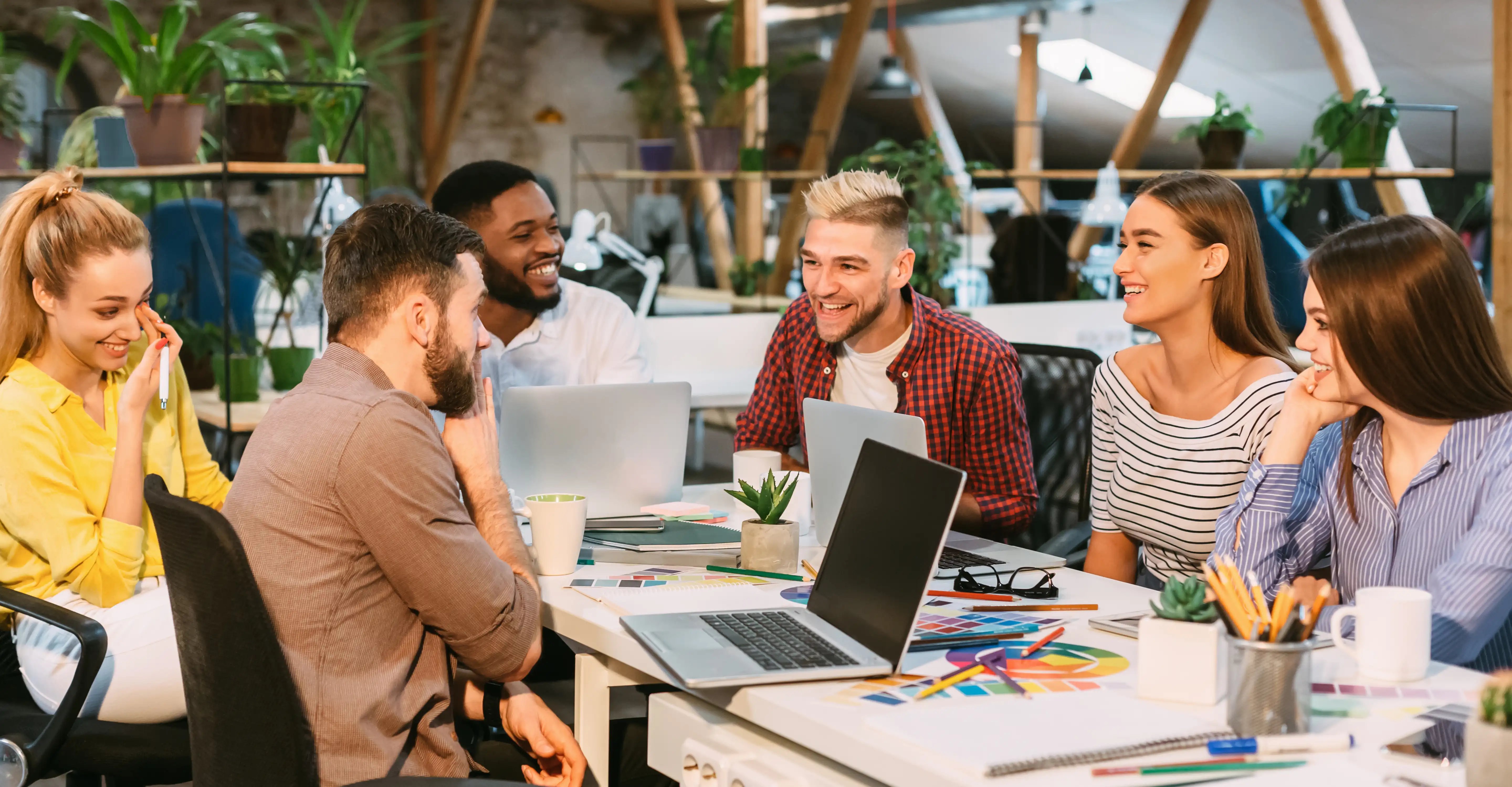 A diverse group of people collaborate in a very friendly way around a table filled with laptops, notebooks and plants in a bright, modern workspace.