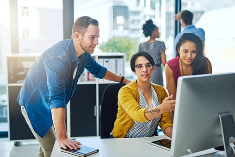 Three people are gathered around a computer screen talking about digital support in a modern office, working together, while two others collaborate in the background.