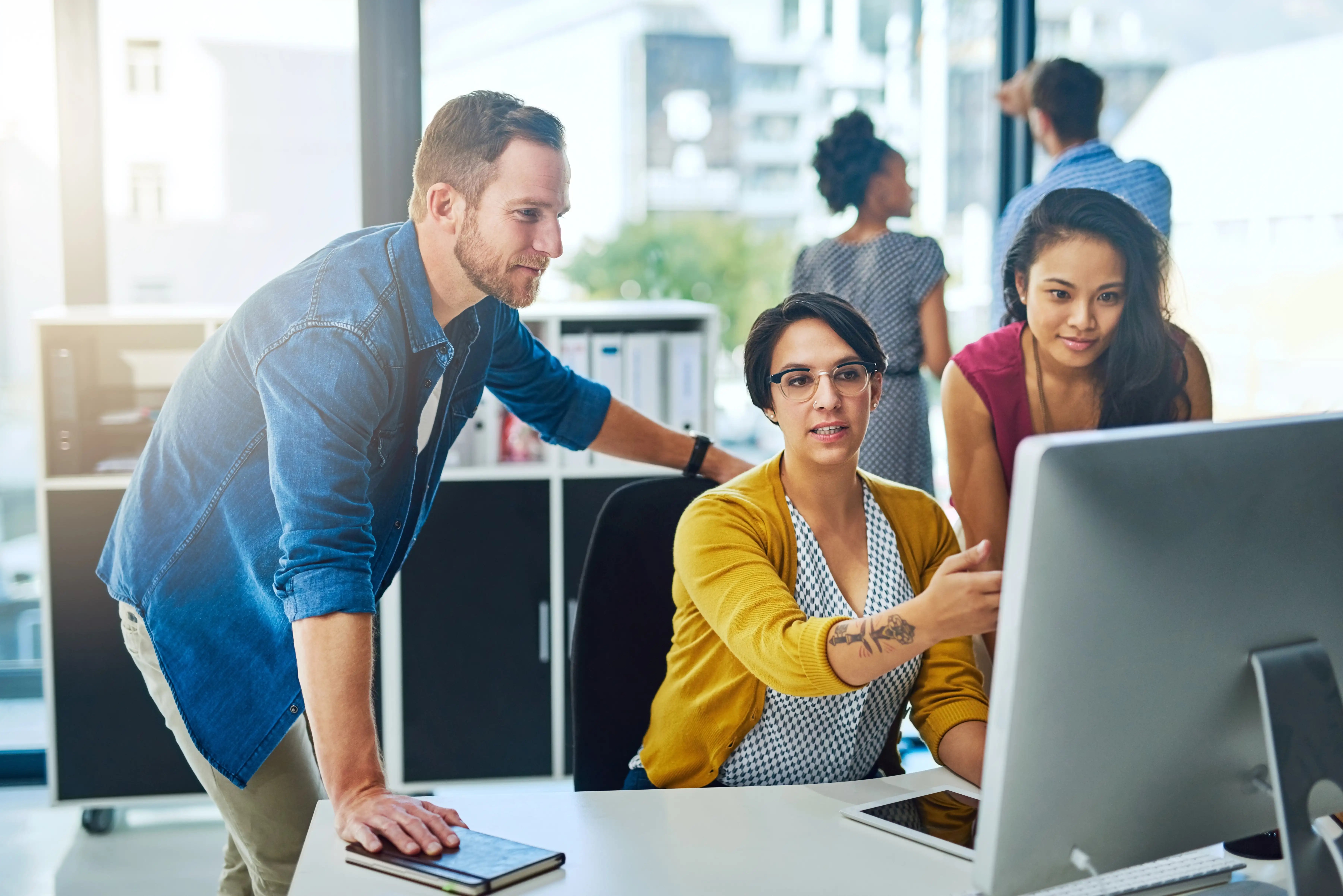 Three people are gathered around a computer screen in a modern office, working together, while two others collaborate in the background.