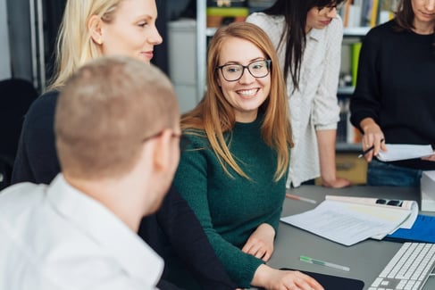 A group of people are sitting and standing around a desk with papers, a keyboard and a computer, following a formation. One person smiles at another.