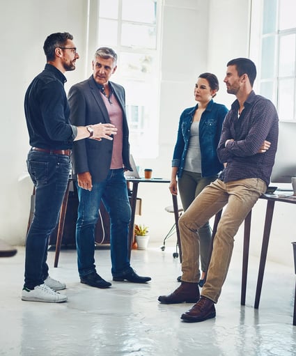 A professional group discussion in a bright office, with four people engaged in conversation near a desk and computer.