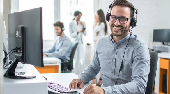 A person wearing headphones sits at a desk with a computer, while colleagues chat in the background in a modern office setting.