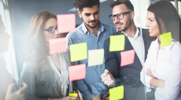 A group of professionals discussing ideas, surrounded by colorful sticky notes on a glass wall in a modern office space.