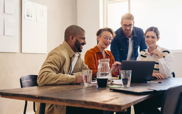 A group of four is gathered around a laptop on a wooden table, engaged in a discussion in a bright, modern office.
