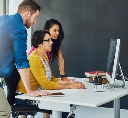Un groupe de trois personnes collabore autour d'un bureau avec un ordinateur, des cahiers et des livres dans un environnement de bureau moderne.