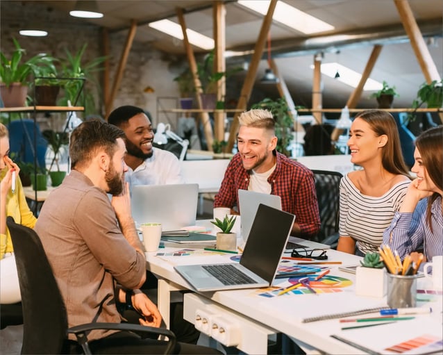 A diverse group of people collaborate in a very friendly way around a table filled with laptops, notebooks and plants in a bright, modern workspace.