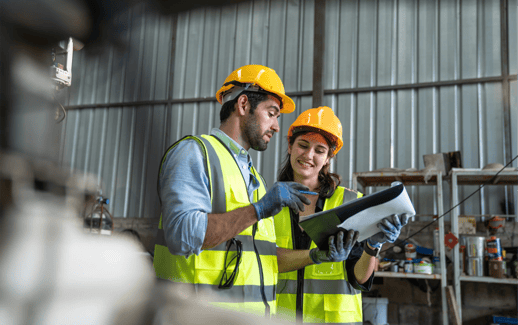 Two construction workers in safety gear discuss plans in a warehouse, surrounded by tools and materials.
