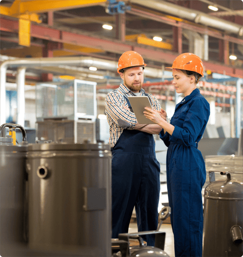Two workers in safety gear at a factory are holding a tablet and talking.