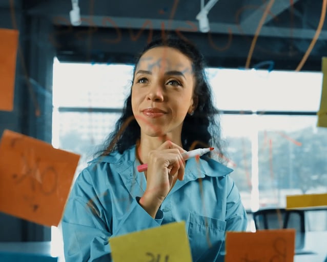 A person in a blue shirt holds a pink marker, pondering in front of colorful sticky notes on a glass board in a modern office.