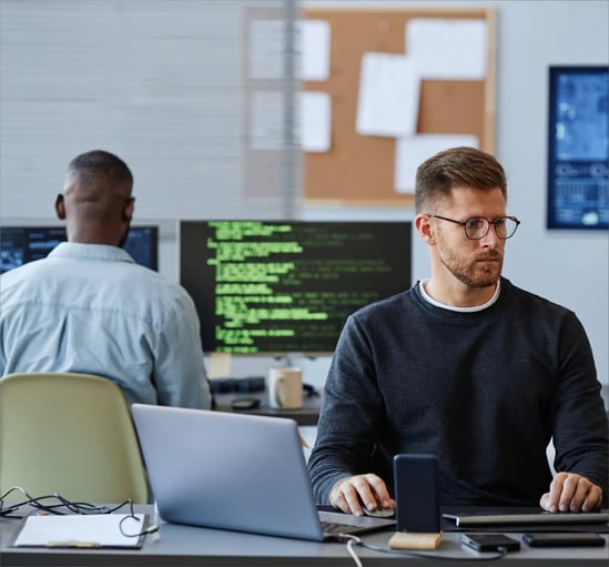 Two people work at a modern desk with computers displaying codes and data, surrounded by office supplies and a notice board.