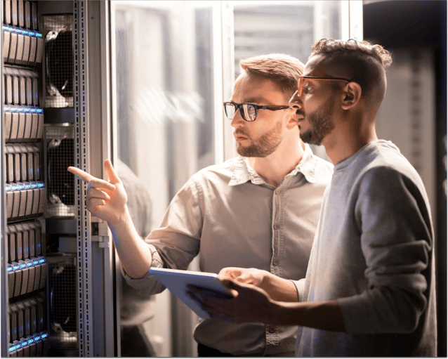 Two people discuss technology in a server room, pointing at equipment while holding a tablet, surrounded by racks of servers.