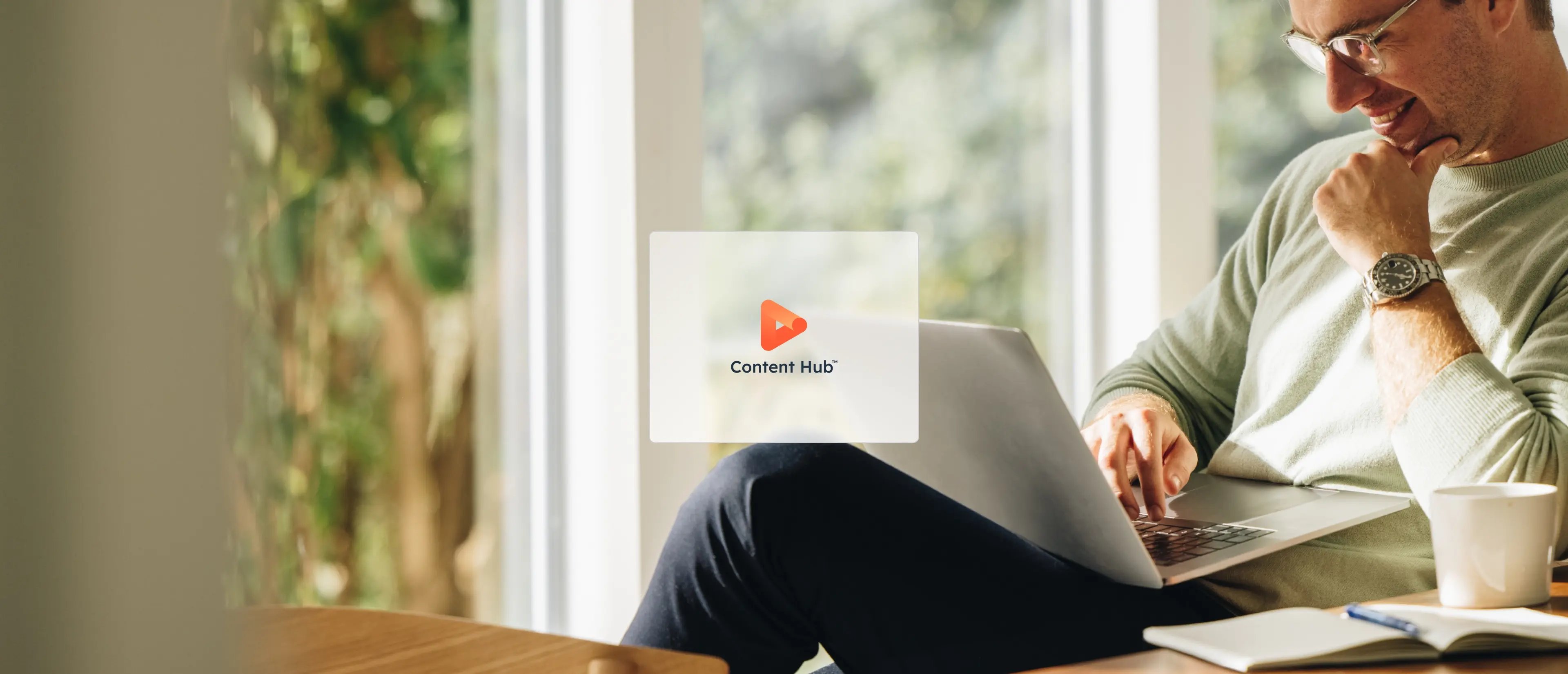 One HubSpot hub icon: Content Hub, is displayed over an image of a person sitting at a wooden table, working on a laptop surrounded by greenery and natural light.