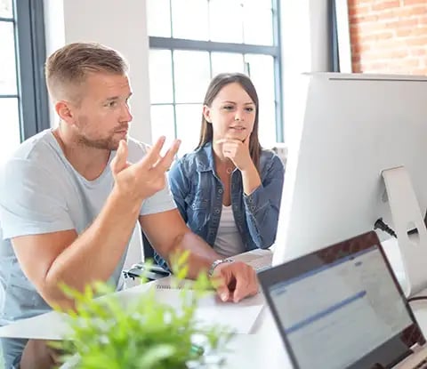 A group discusses ideas in a bright, modern workspace, with a large screen, laptops and plants on the table.