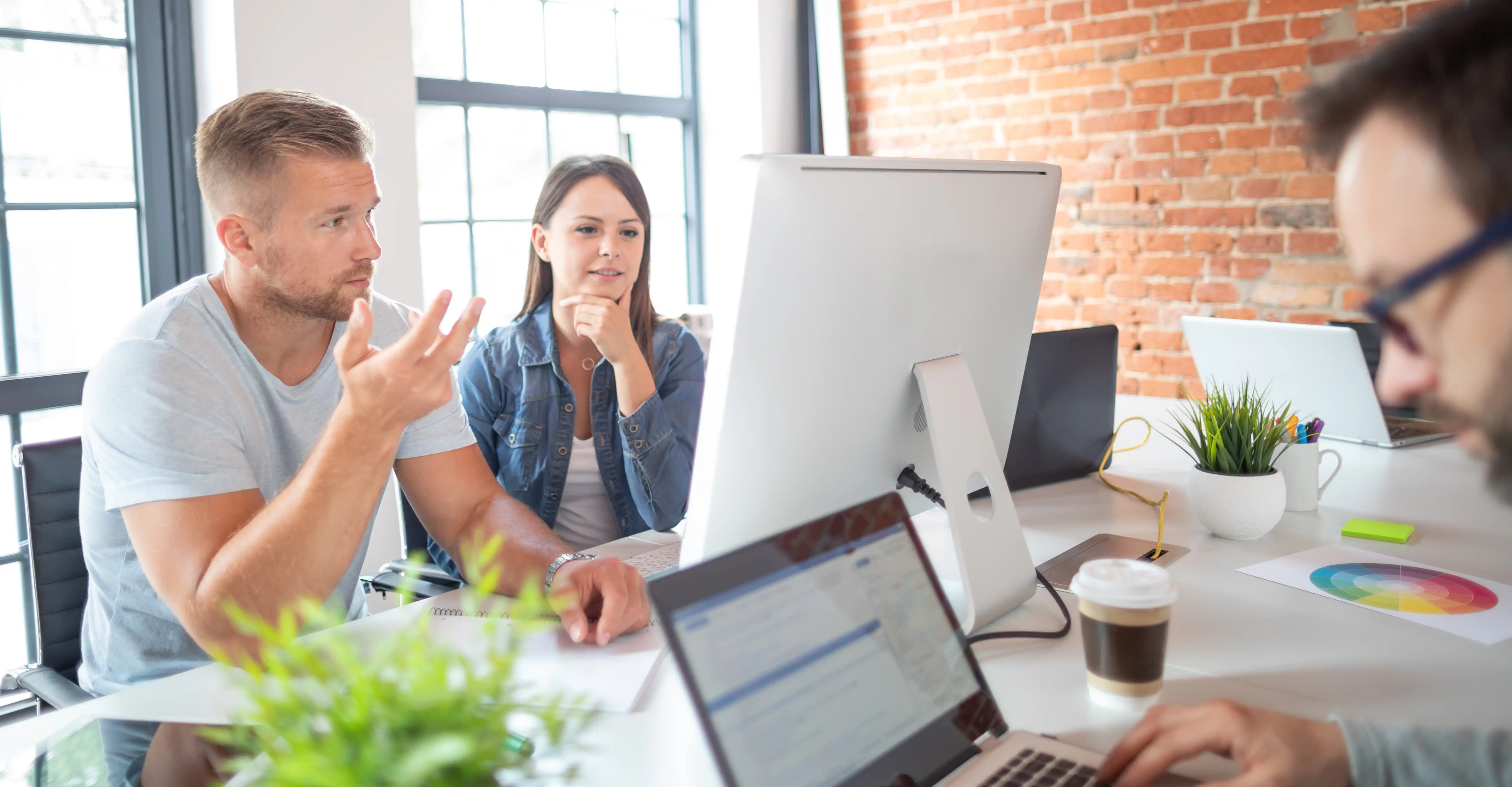 A group discusses ideas in a bright, modern workspace, with a large screen, laptops and plants on the table.