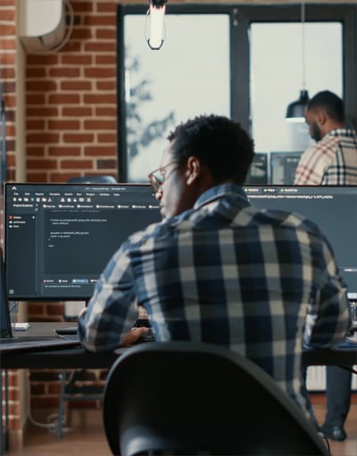 A person in a checkered shirt sits at a desk, focusing on dual monitors displaying code in a modern, brick-walled office.