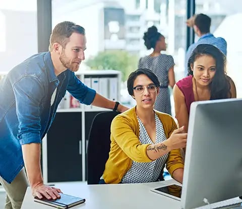 Trois personnes sont réunies autour d'un écran d'ordinateur dans un bureau moderne, travaillant ensemble, tandis que deux autres personnes collaborent à l'arrière-plan.