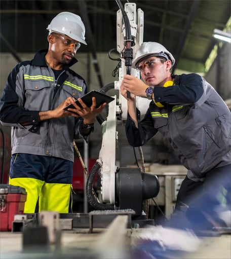 Two engineers in safety gear review a tablet while working on a robotic arm in an industrial setting.