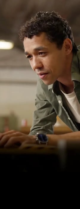 A person leans over a table, focused on a laptop, wearing a green shirt and a wristwatch, in a bright industrial workspace.